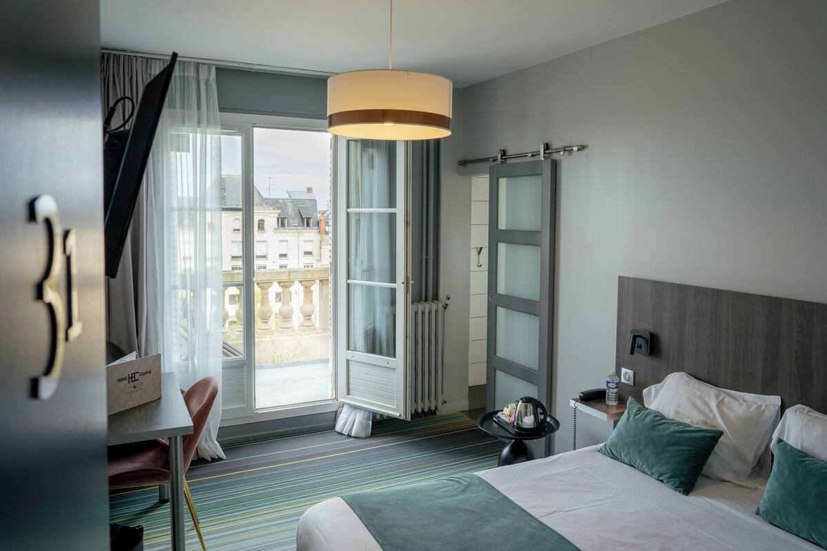 A hotel room with a bed, a desk, a chair, a window with curtains, a radiator, and a view of buildings outside.