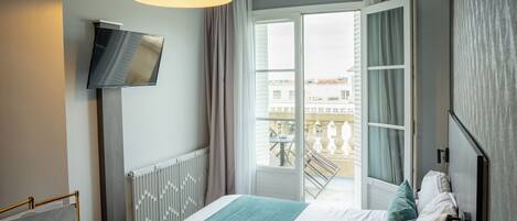Una habitación de hotel con cama, televisor montado en la pared, balcón con vistas a edificios y un cartel con el nombre del hotel.