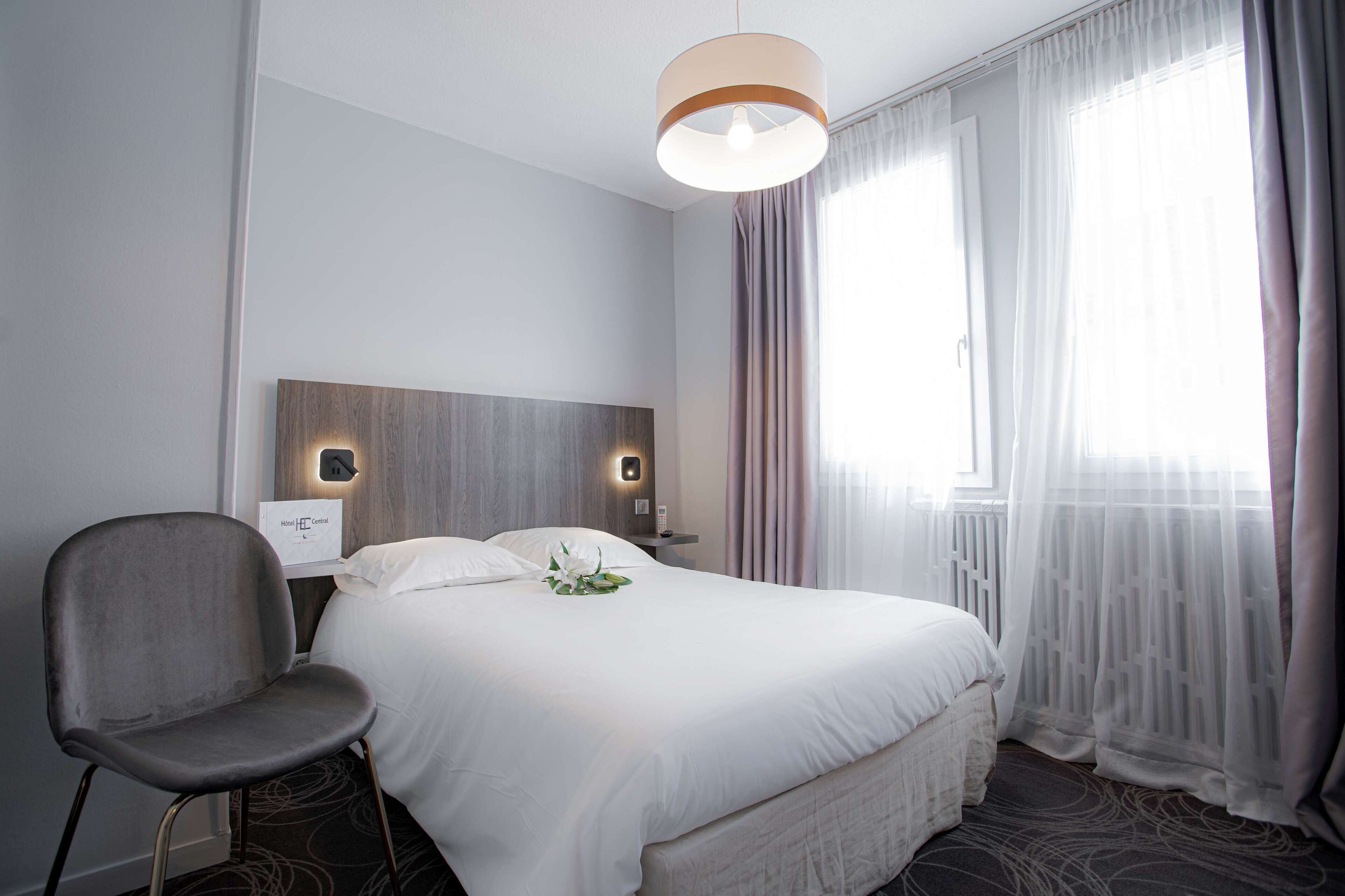 Une chambre d’hôtel avec un grand lit, une chaise, une table de chevet avec une lampe et une fenêtre avec des rideaux légers.