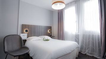 Une chambre d’hôtel avec un grand lit, une chaise, une table de chevet avec une lampe et une fenêtre avec des rideaux légers.