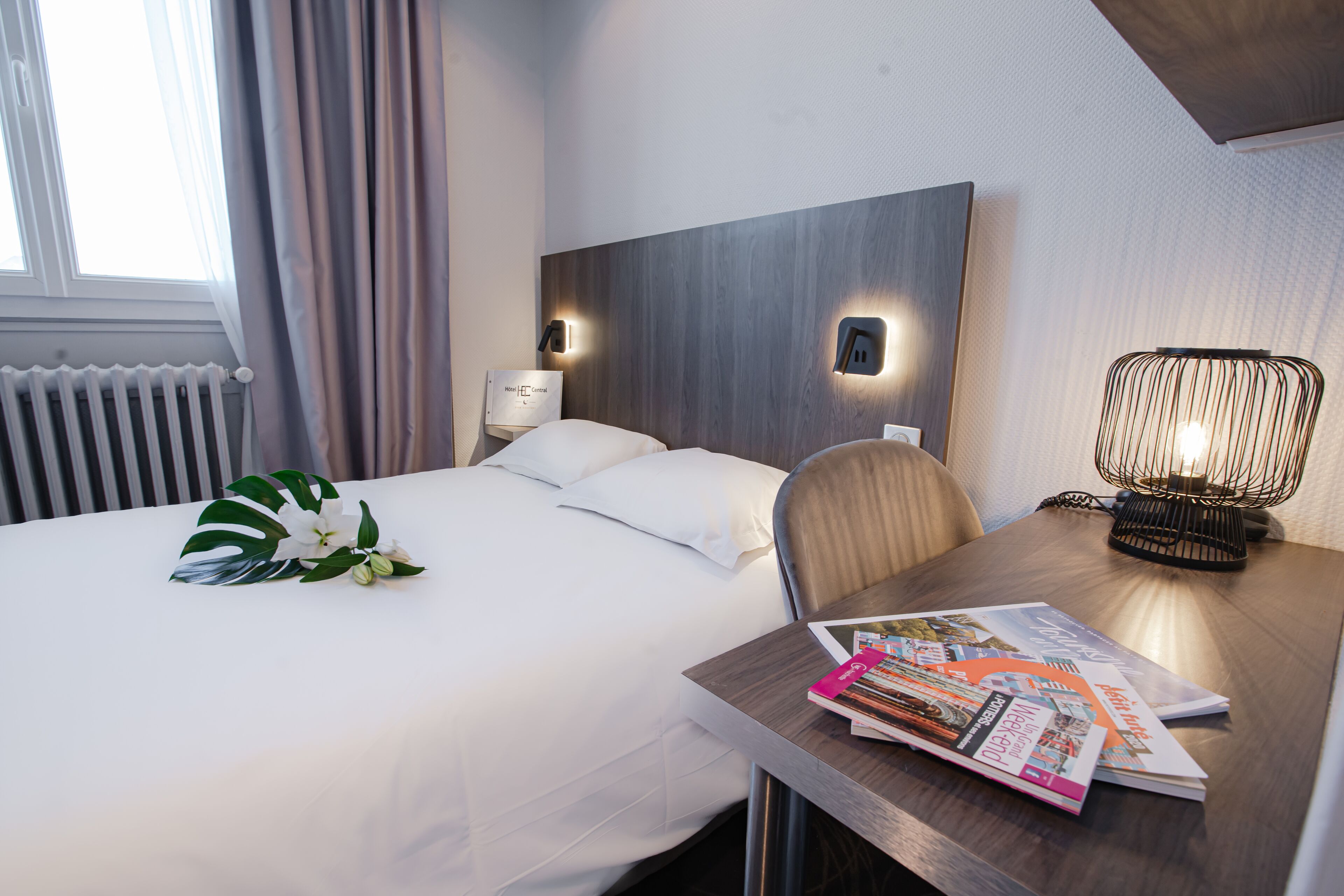 A hotel room with a neatly made bed, a wooden headboard, a bedside table with a lamp, and a window with curtains.