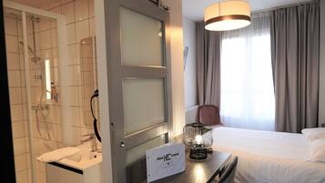 A hotel room with a shower, a bed, a bedside table with a sign, and a window with curtains.
