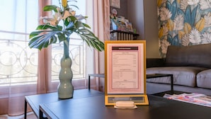 A menu board with a list of food and drink items, a vase with a plant, and a sofa in a room with a window and curtains. - Hotel Central (Poitiers)