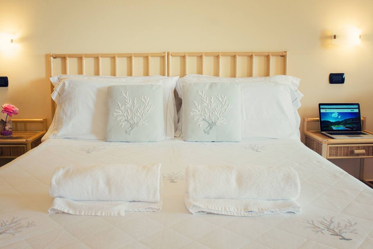 Een net opgemaakt bed met witte lakens, twee kussens met geborduurde motieven, en een laptop op een nachtkastje.