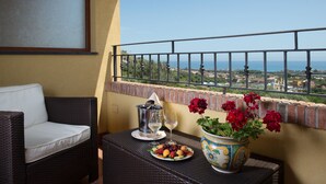 Een balkon met een rieten stoel, een tafel met een fruitschaal, een wijnglas, een metalen bak en een bloempot.