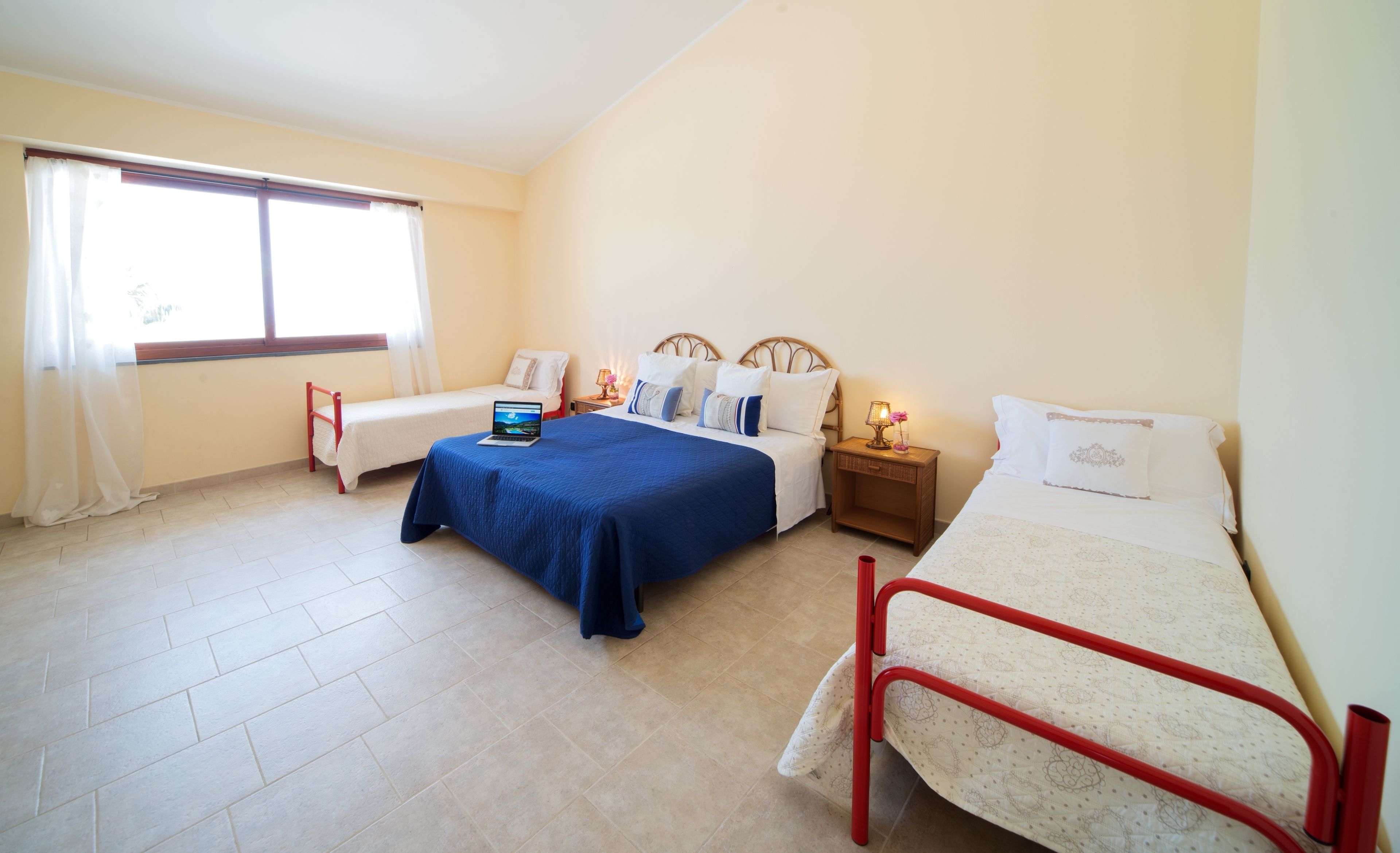 Una habitación con dos camas, una ventana con cortinas, un ordenador portátil sobre una cama y una mesita de noche con una lámpara.