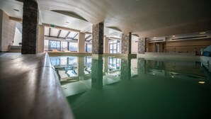Une vaste piscine intérieure à l’eau claire, agrémentée de piliers en pierre et de grandes fenêtres qui laissent entrer la lumière naturelle.