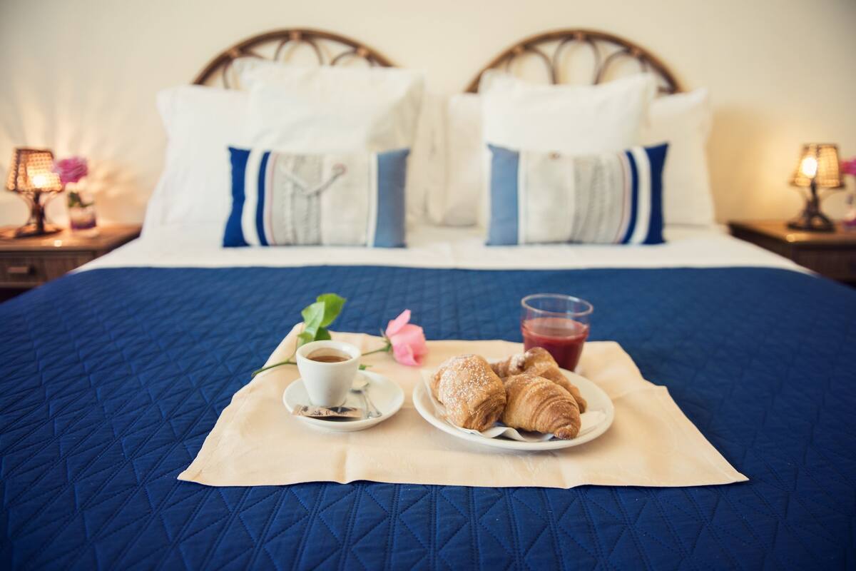 Een netjes opgemaakt bed met een blauwe quilt, een bord croissants, een kop koffie en een glas sap op een beige servet.