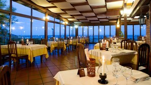 Un restaurant avec de nombreuses tables dressées pour le service, de grandes fenêtres offrant une vue sur la mer et un sol carrelé.