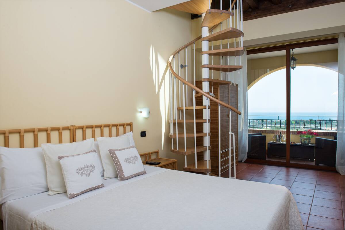 Een slaapkamer met een groot bed, een houten hoofdbord, een wenteltrap en een balkon met uitzicht.