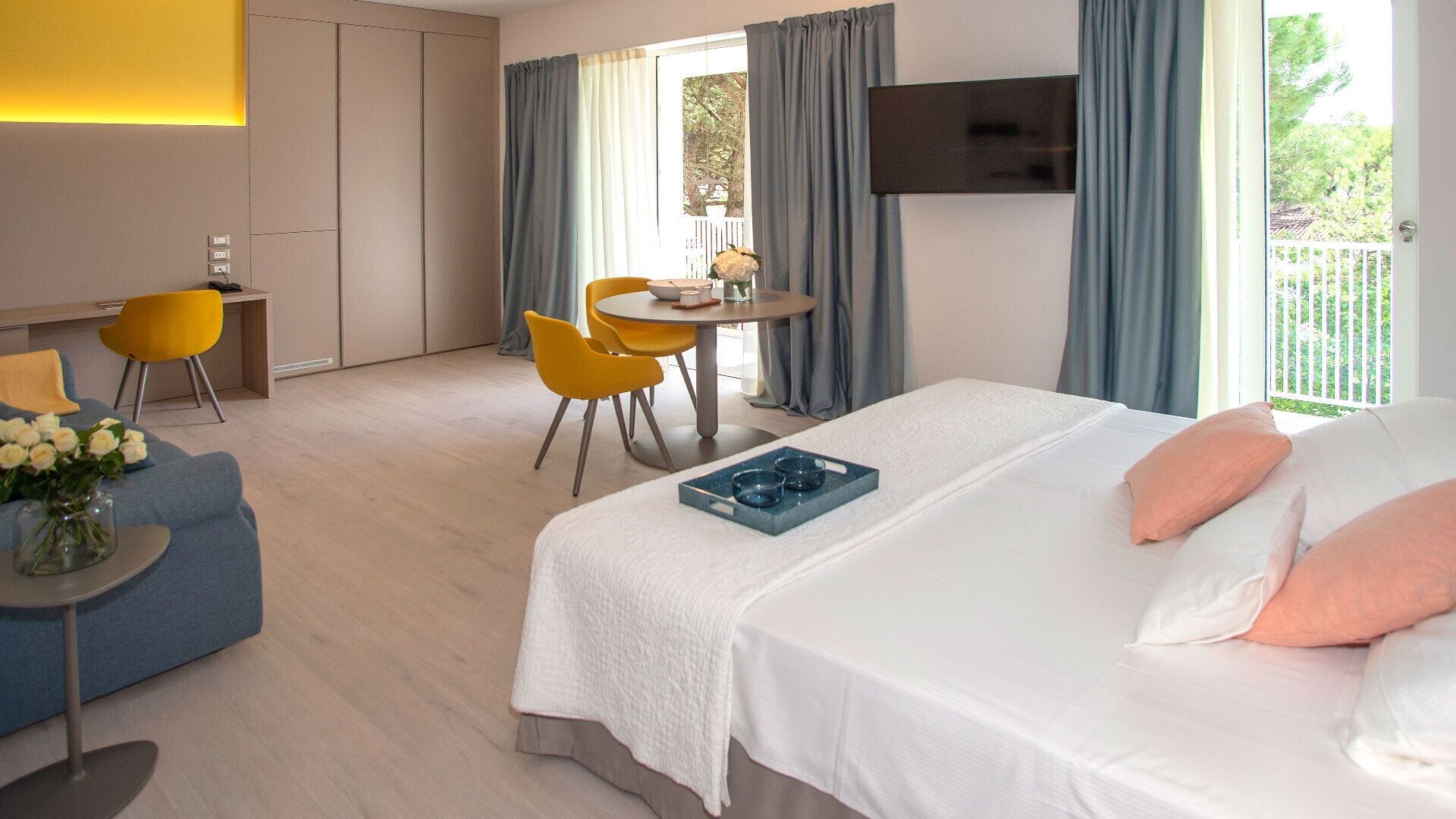 Habitación de hotel con una cama grande, un escritorio con silla, una mesita con otra silla y un televisor montado en la pared.