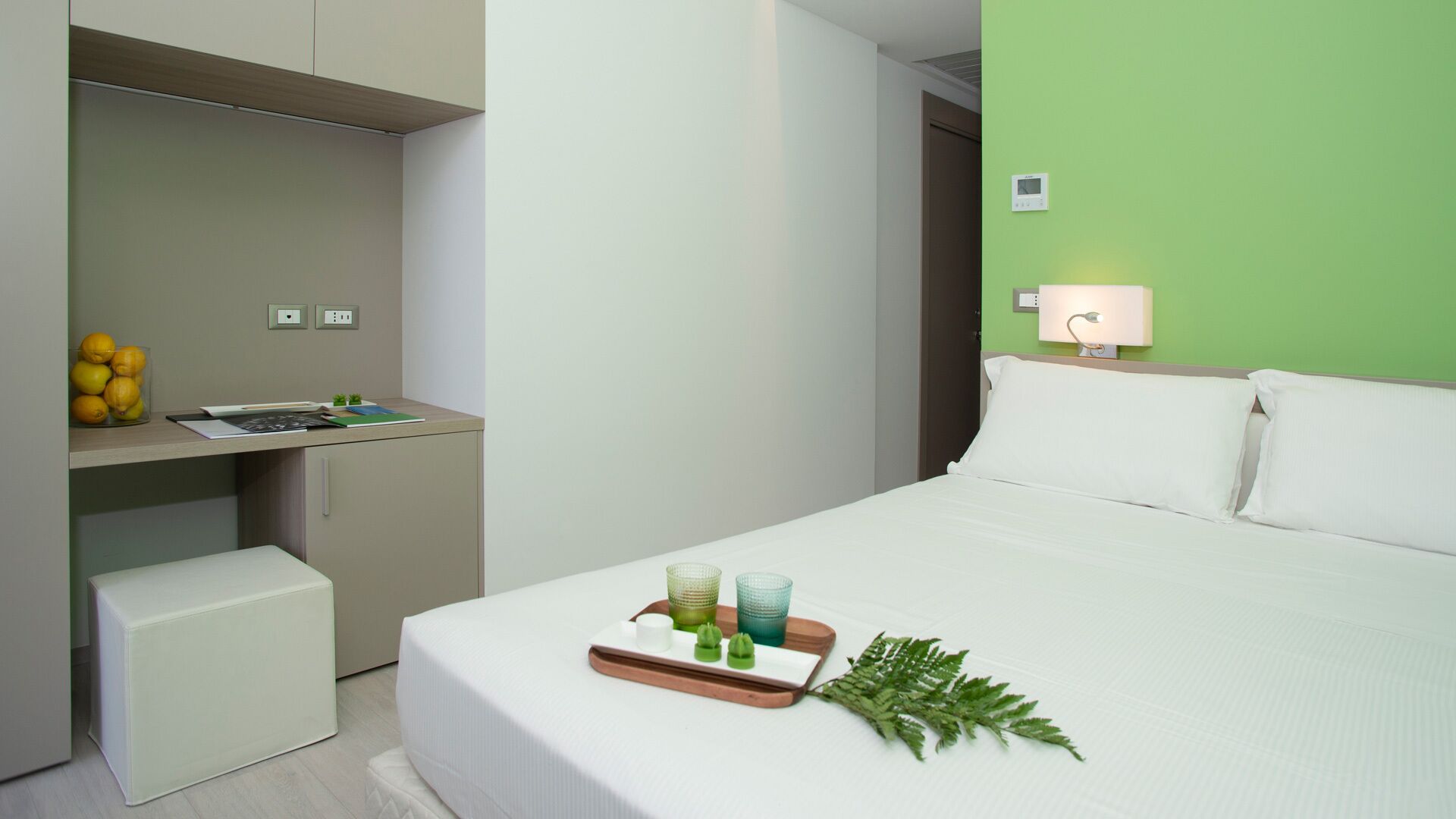 A modern hotel room with a bed, a desk, and a small kitchenette.