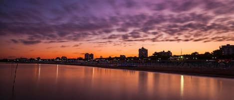 Paisagem urbana ao entardecer, com edifícios ao longo da orla marítima, água calma refletindo o céu e uma praia com guarda-sóis.