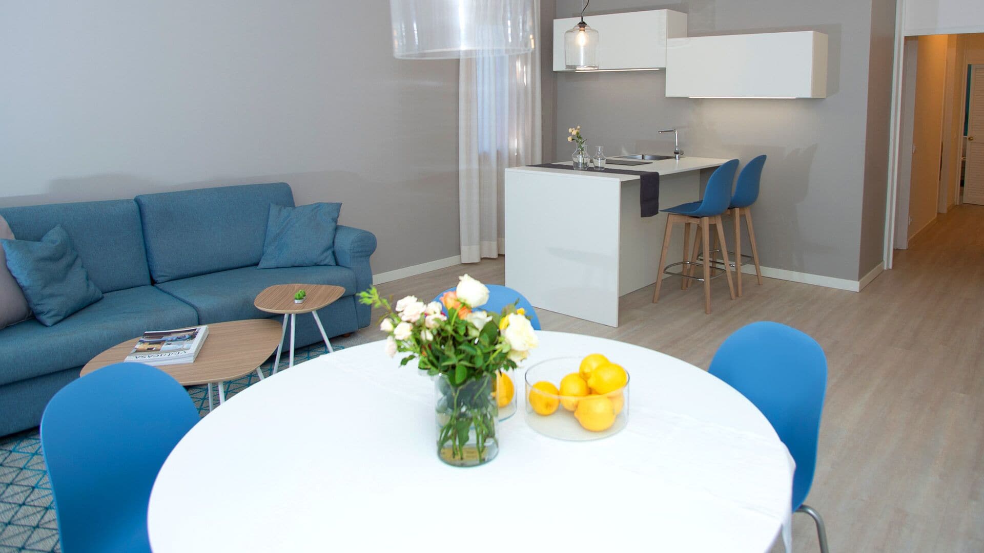 Ein modernes Wohnzimmer mit einem blauen Sofa, einem kleinen Couchtisch und einem Essbereich mit einem weißen Tisch und blauen Stühlen.