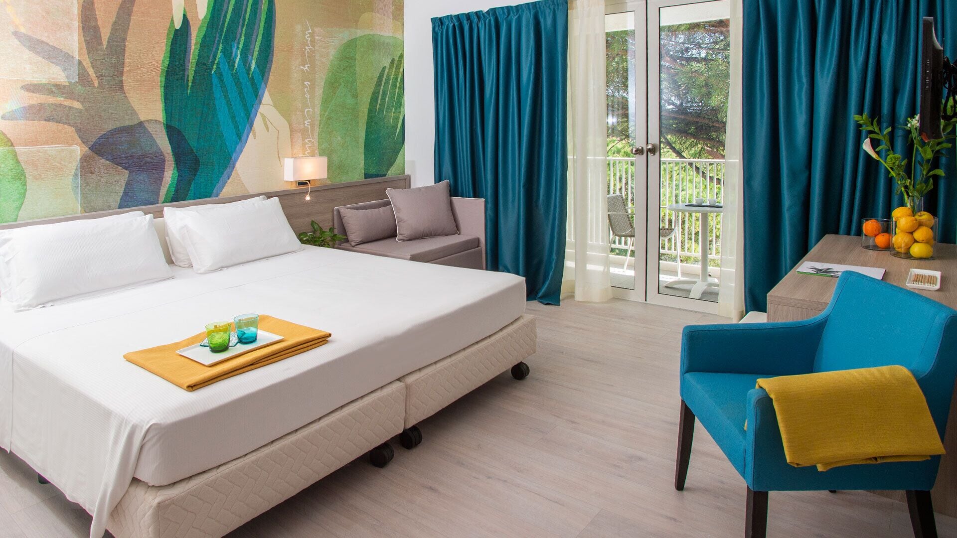 Een hotelkamer met een bed, een stoel, een tafel met fruit en uitzicht op groen door de gordijnen.