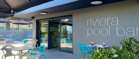 Zona de bar junto a una piscina exterior con sillas y mesas turquesa, una entrada con puerta de vidrio y un letrero que dice "rivier pool bar".