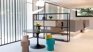 Un interior moderno con una mesa redonda, dos taburetes, un estante decorativo con flores y vistas a la vegetación a través de una ventana.