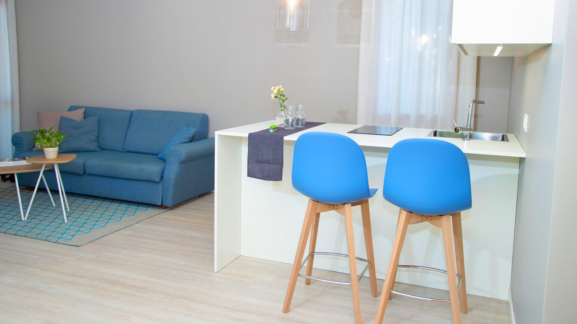 Un salon moderne avec un canapé bleu, une petite table basse et une plante en pot. On y trouve également un coin cuisine avec un comptoir blanc, deux tabourets de bar bleus et un évier.