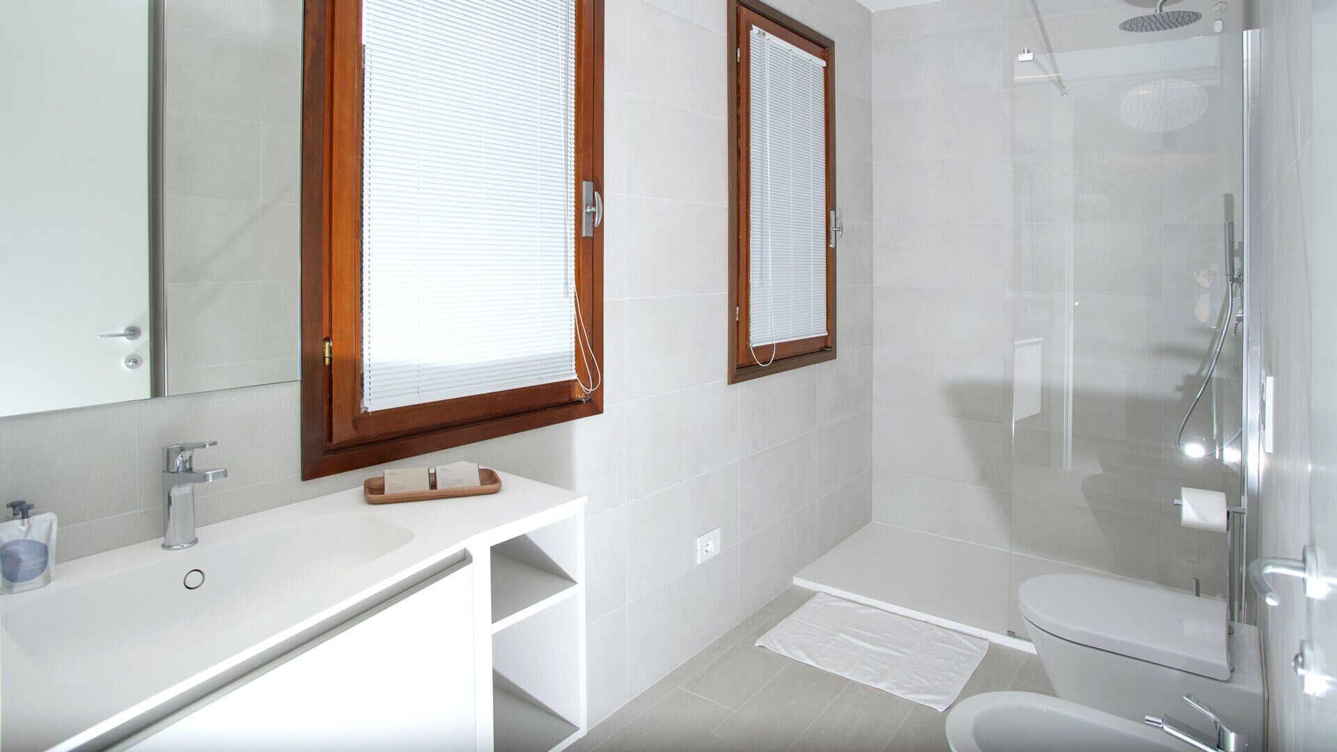 Et moderne badeværelse med vask, underskab og toilet i hvid. To vinduer med trærammer og persienner, et glasbadeværelsesafskærmning og en hvid badeværelsesmåtte på gulvet.