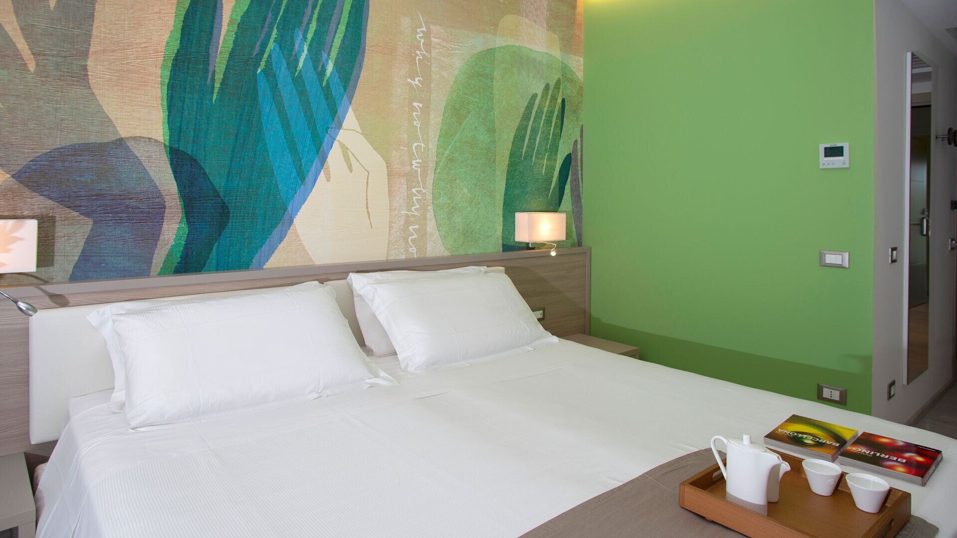 A hotel room with a bed, a nightstand, a tray with cups and a book, and a green wall.