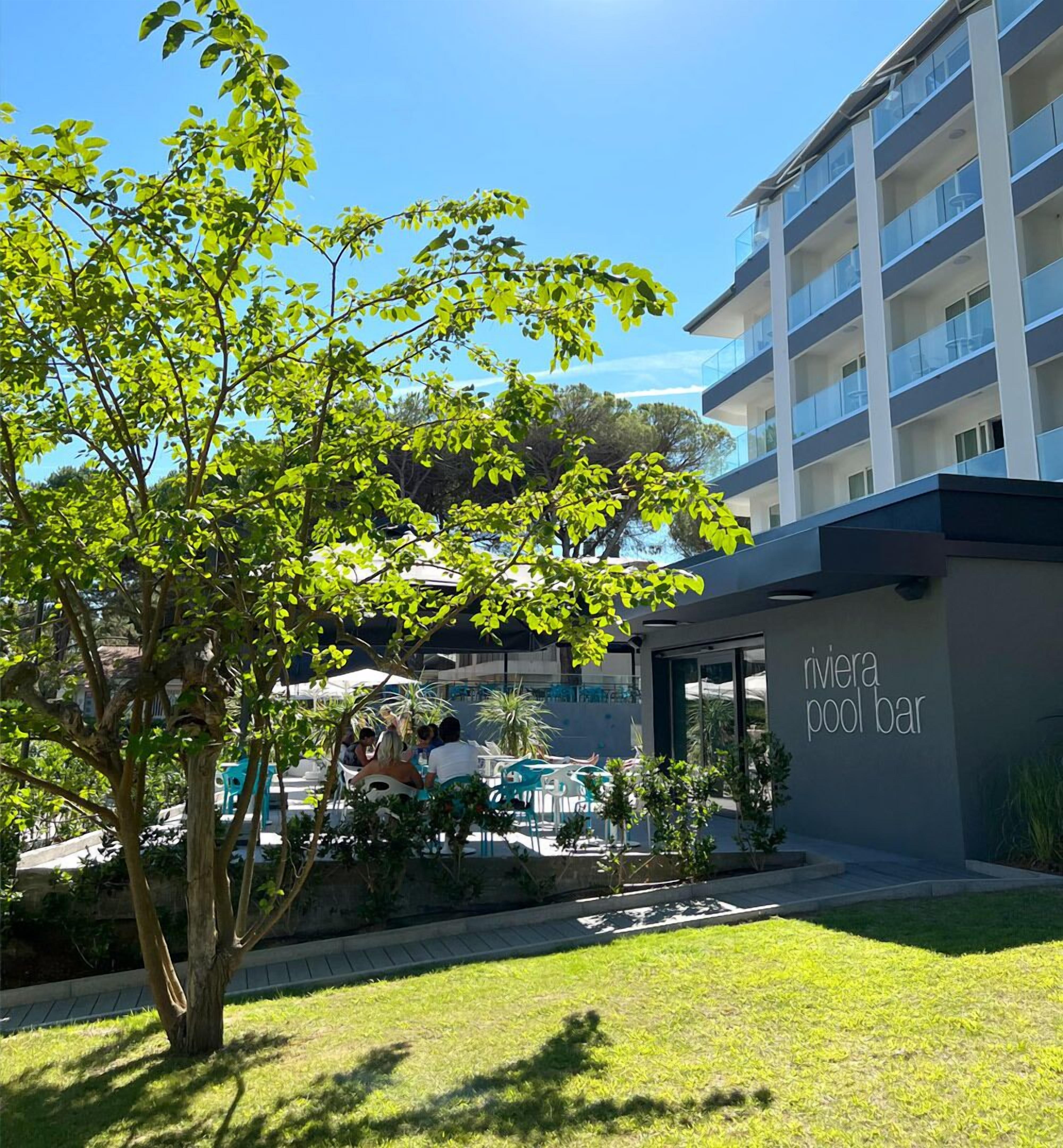 A modern building with a pool bar area, outdoor seating, and a tree in the foreground.