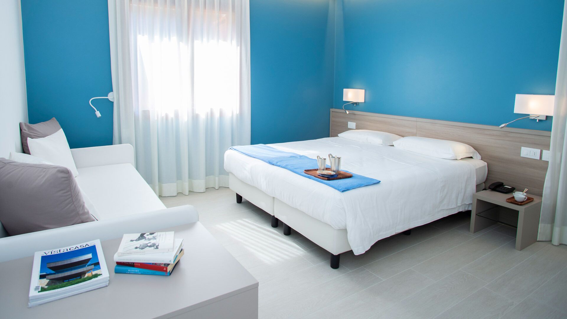 Una camera d'albergo moderna con un letto grande, comodini, un divano bianco e una finestra con tende leggere.