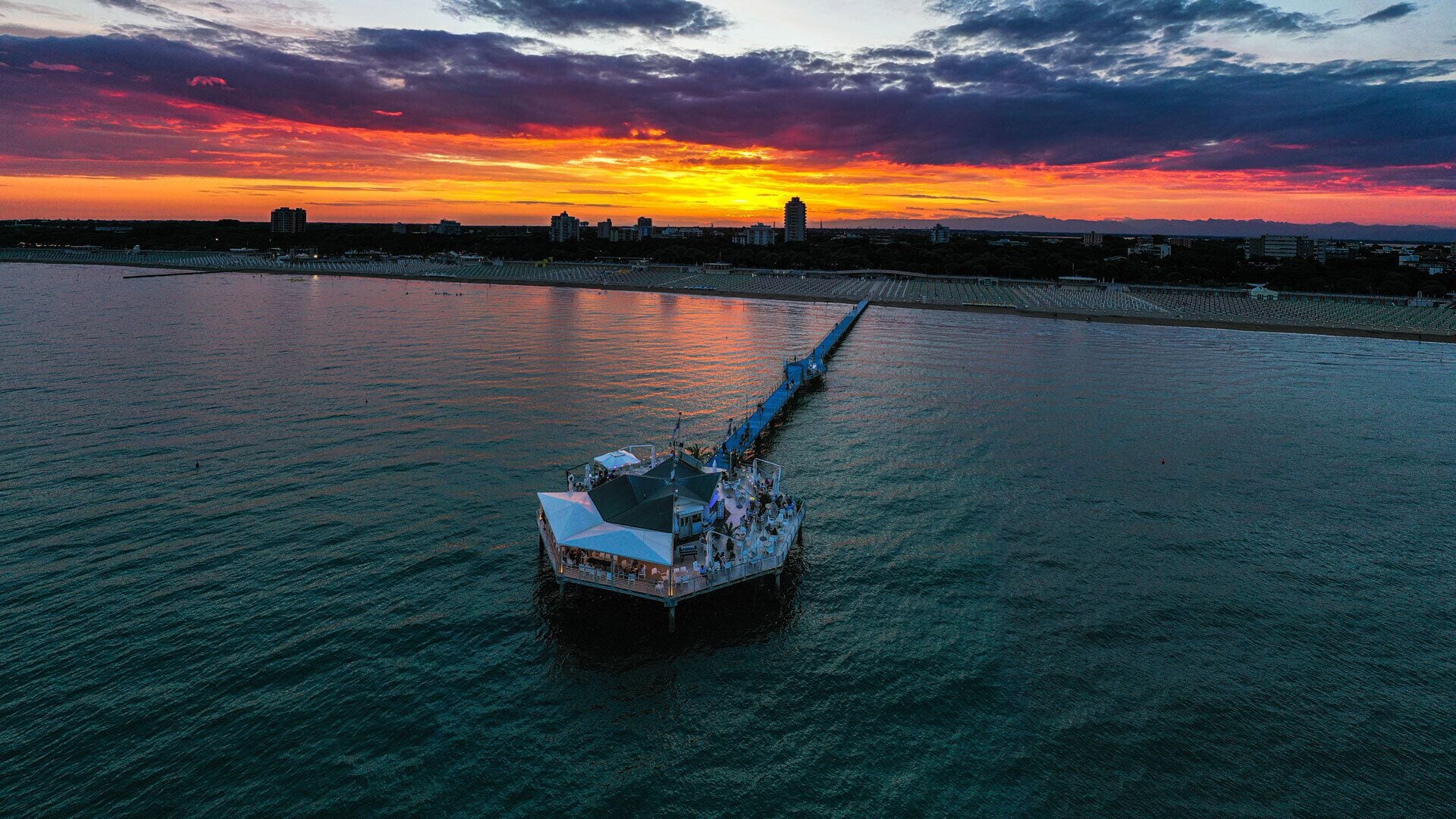 A waterfront boat with a covered deck and seating, docked at a pier with a sunset in the background.