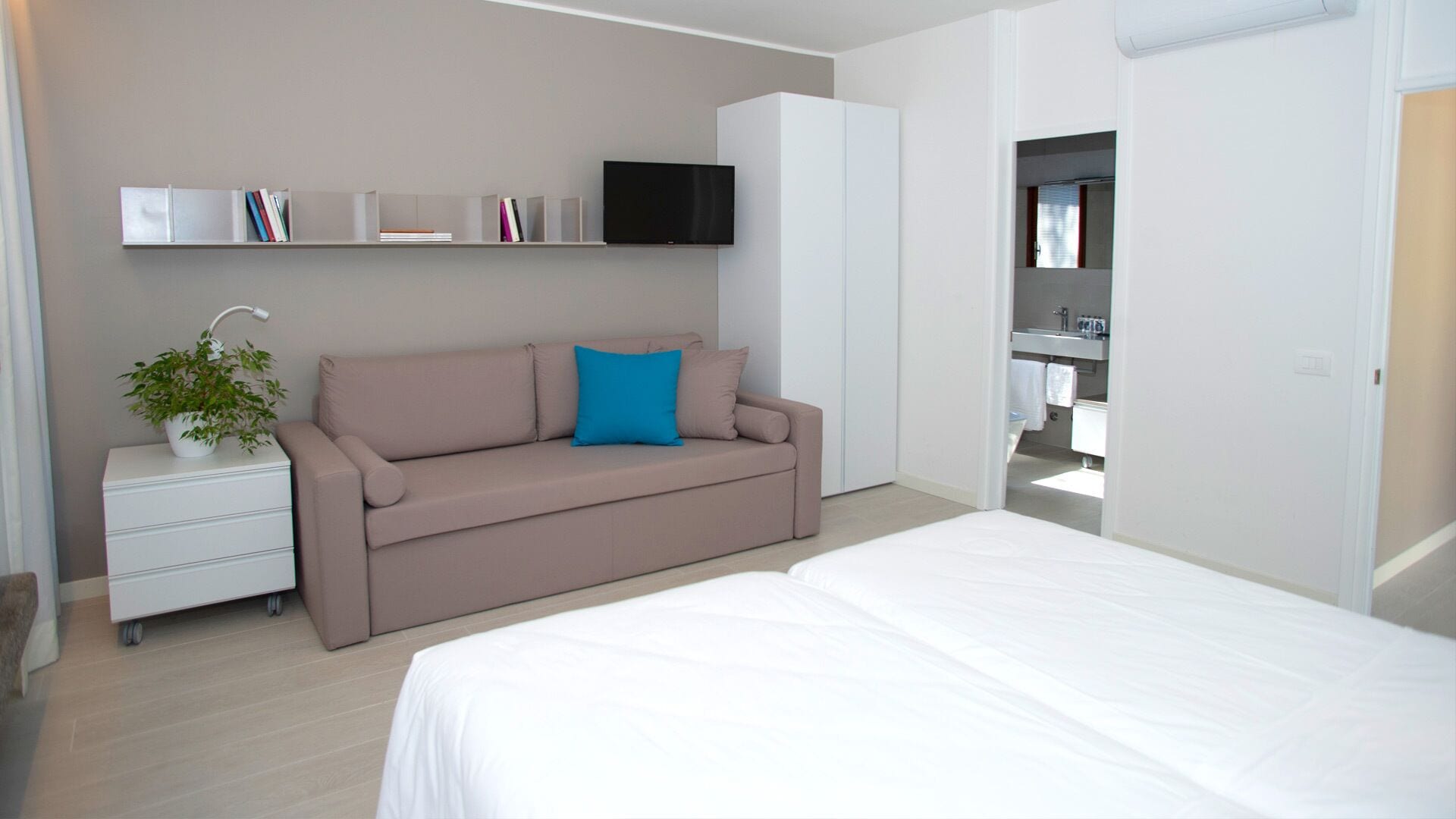 A modern bedroom with a beige sofa, a white bedside table, a flat-screen TV mounted on the wall, and a visible bathroom with a sink and mirror.