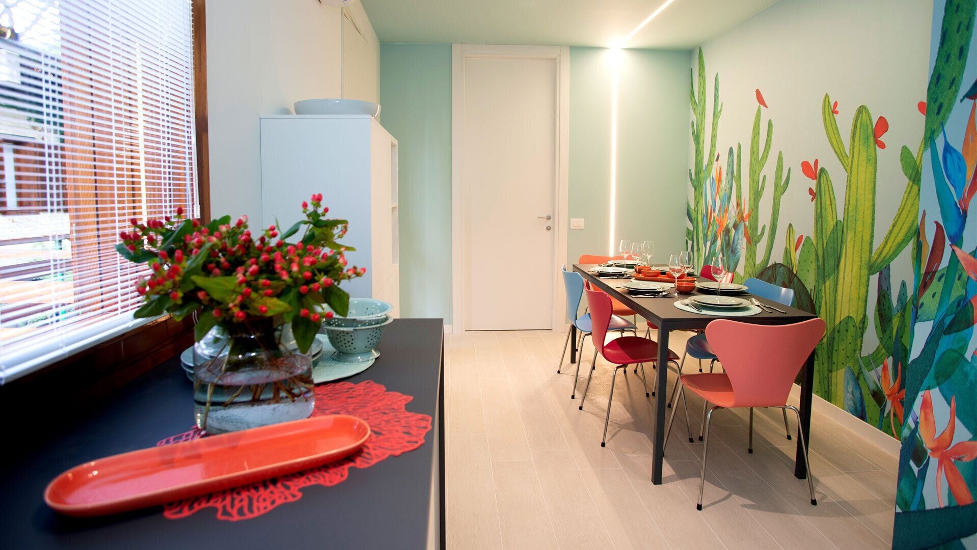 Un coin repas avec une table dressée pour quatre personnes, des chaises aux couleurs vives, un vase rempli de baies rouges et une fresque murale sur le thème des cactus.