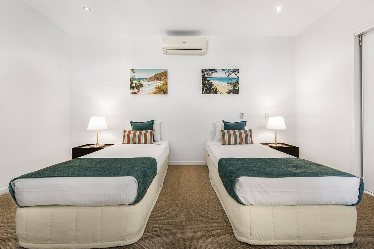Habitación de hotel con dos camas, cada una con una manta verde y almohadas blancas, una mesita con una lámpara y dos cuadros en la pared que representan escenas de playa.