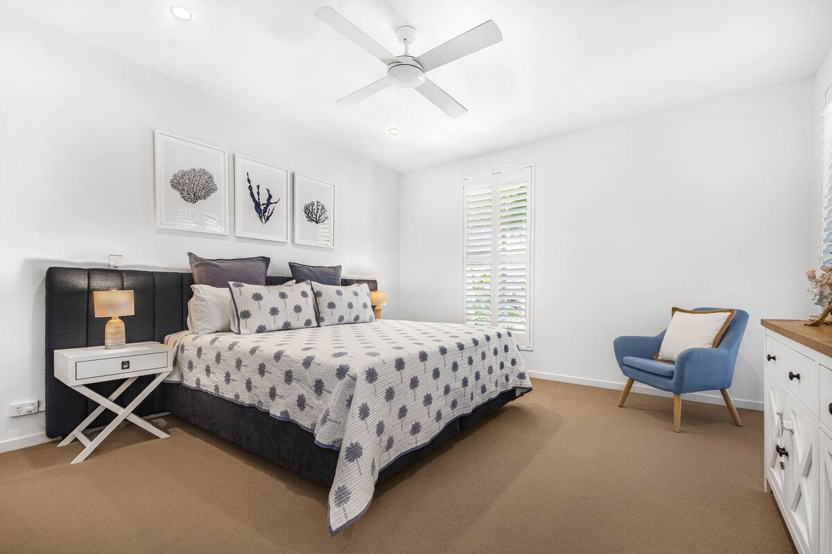 Un dormitorio con cama, mesita de noche, lámpara, sillón y ventilador de techo.