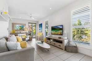 Una sala de estar moderna con sofá, mesa de centro, televisor y ventanas amplias con persianas.