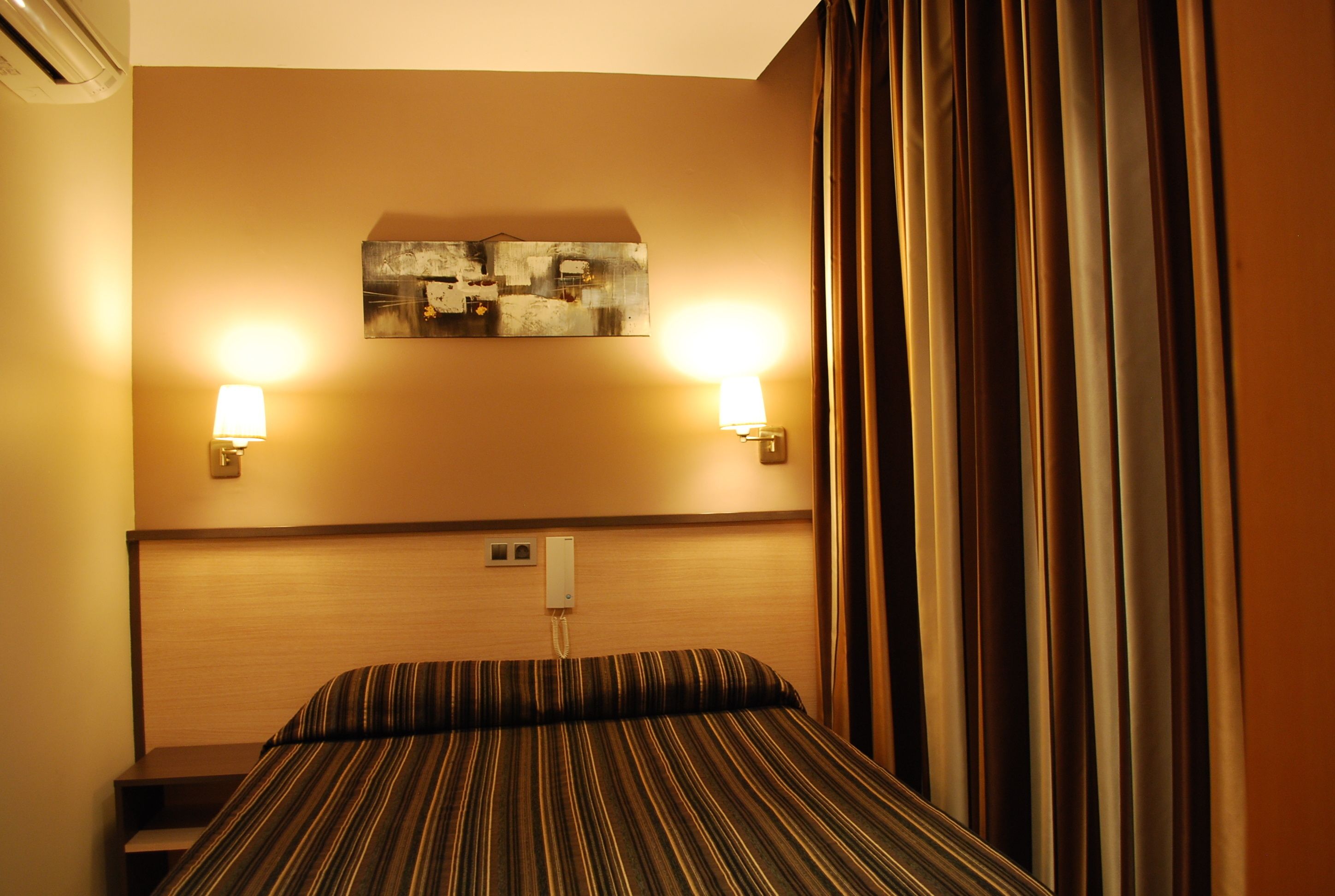 Habitación de hotel con cama, dos lámparas fijadas a la pared, un cuadro sobre la cama y un teléfono en la pared.
