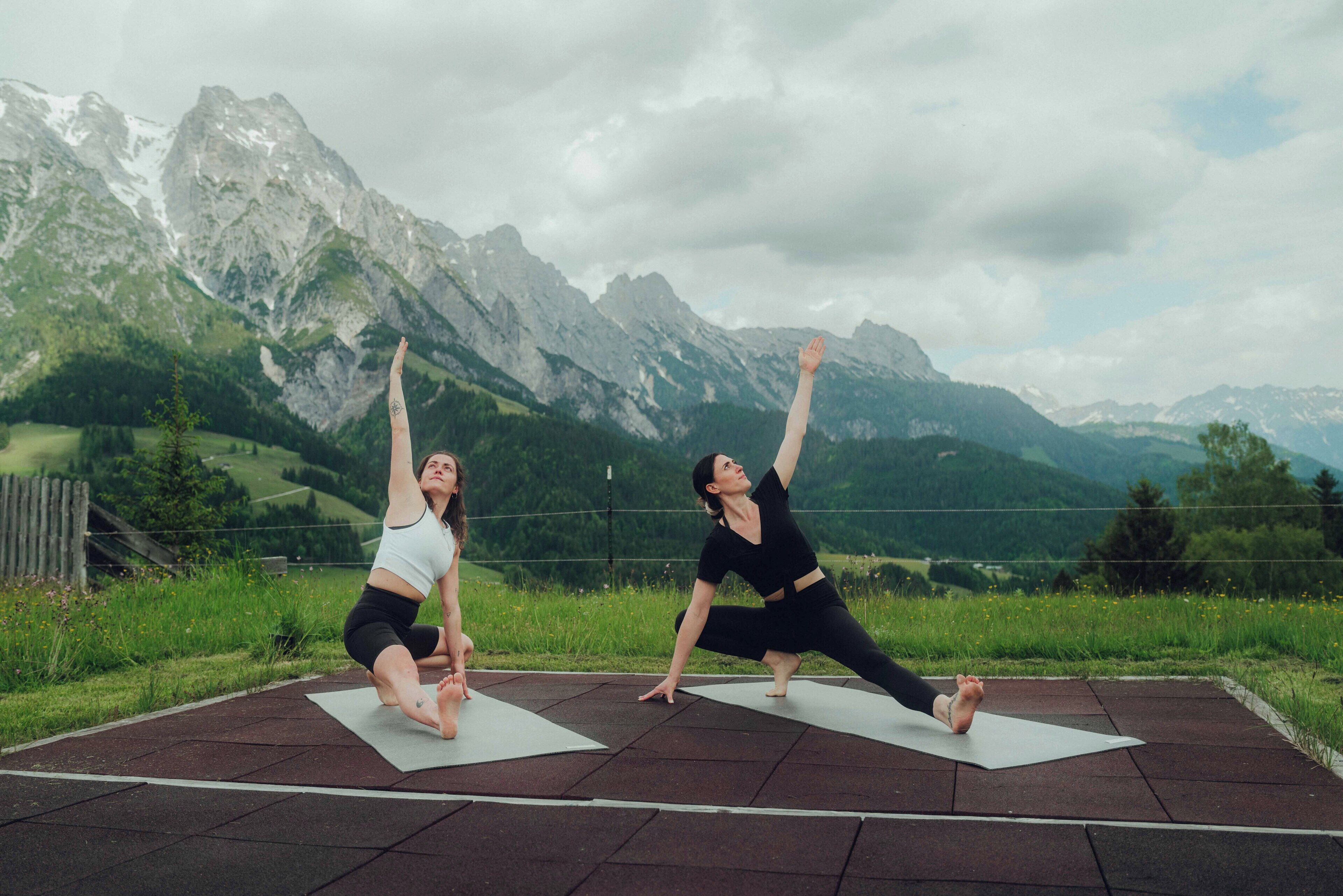 Two people practicing yoga on mats outdoors with a mountainous backdrop.