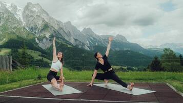 Two people practicing yoga on mats outdoors with a mountainous backdrop.