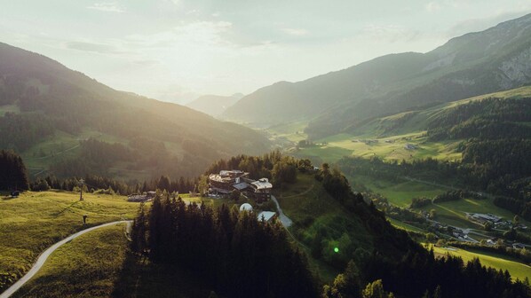 A mountain lodge with a winding road leading to it, surrounded by forested hills and valleys. - Forsthofalm (Leogang)