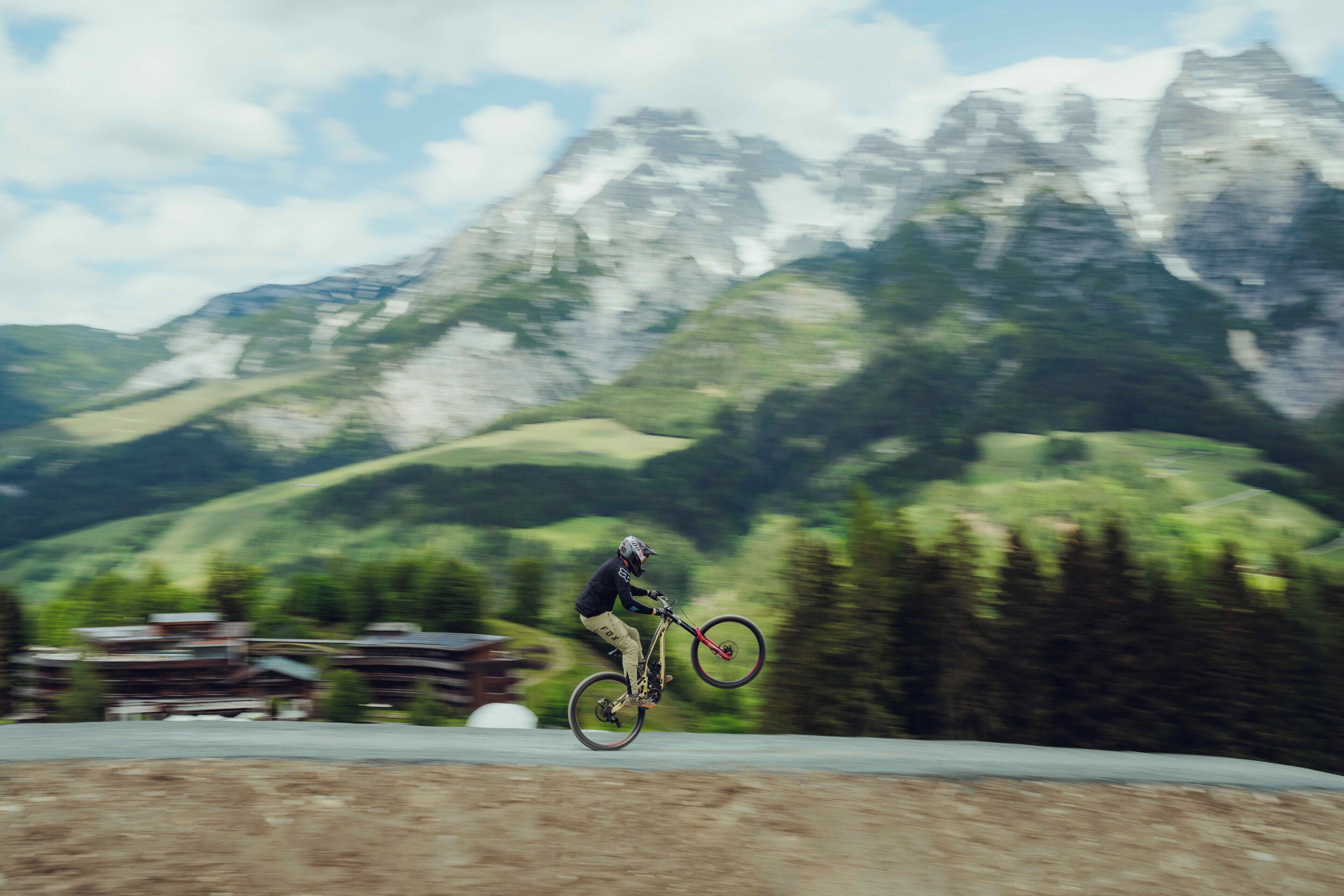 A mountain biker performing a jump on a dirt track with a mountainous backdrop.