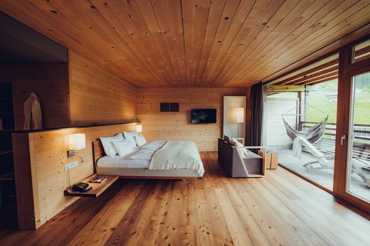 A wooden cabin bedroom with a bed, bedside table, lamp, and a hammock outside the window.