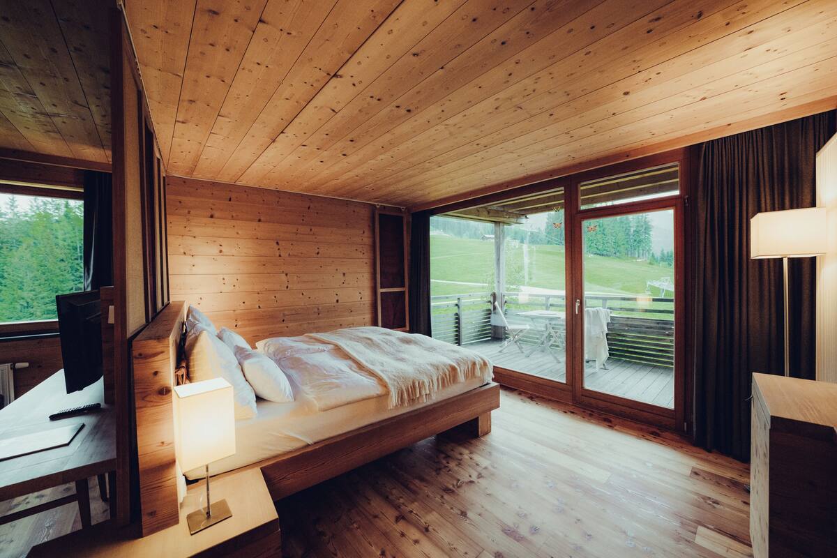 A wooden cabin bedroom with a bed, a desk, and a large window.