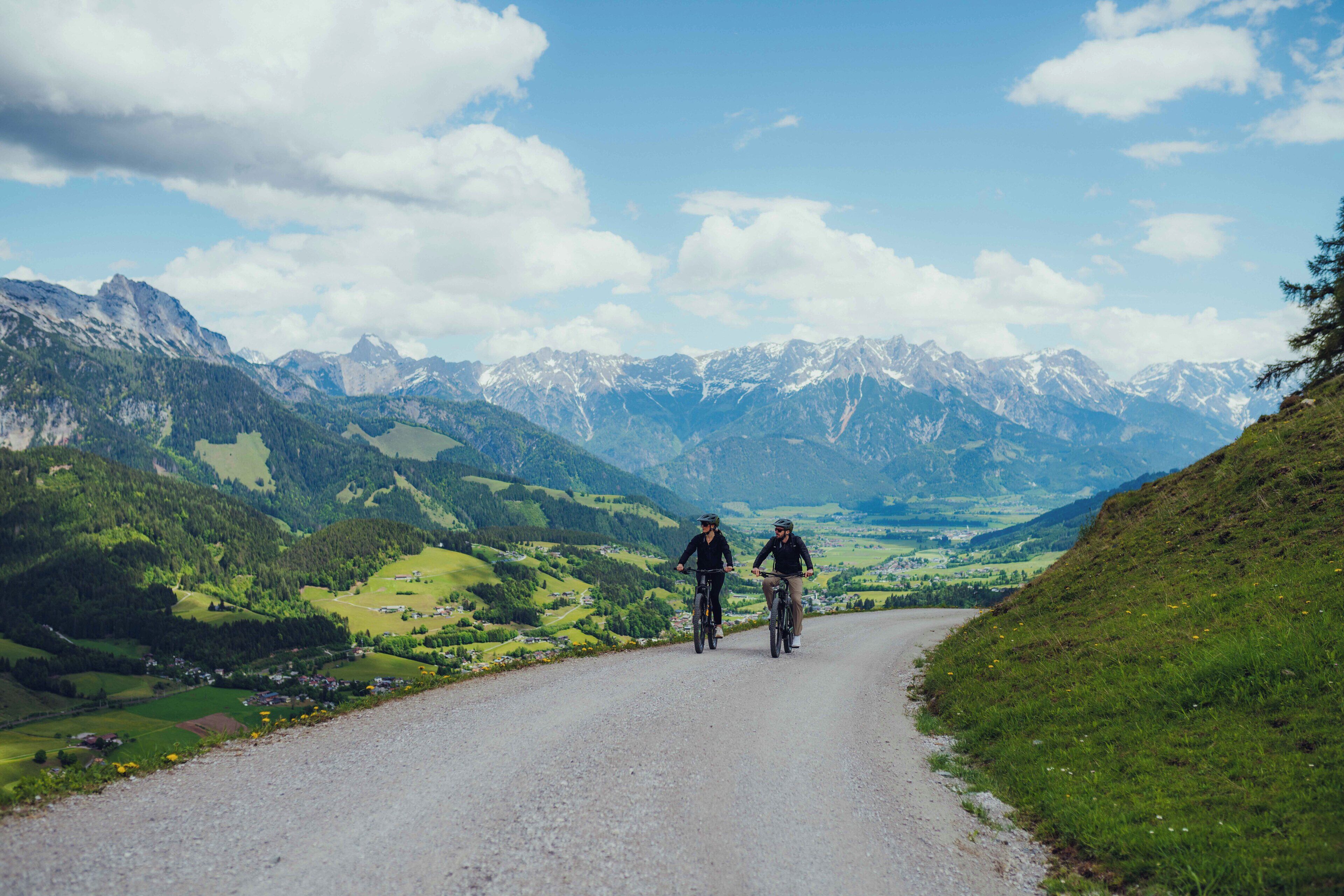 A mountain road with cyclists, surrounded by green hills and distant snow-capped mountains under a partly cloudy sky.