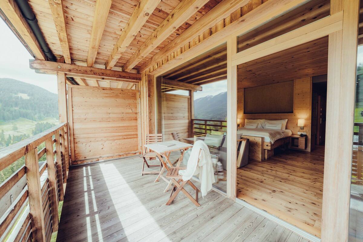 A wooden cabin with a balcony, a dining area, and a bedroom with a bed and nightstands.
