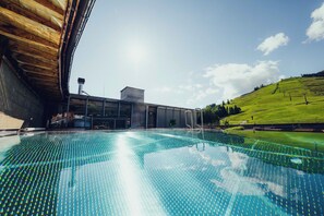 A swimming pool with a clear blue water surface, surrounded by a wooden deck and a modern building with a glass facade. - Forsthofalm (Leogang)