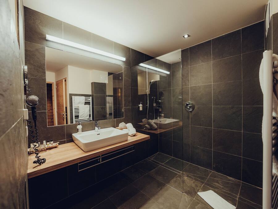 Un baño moderno con un espejo grande, tocador de madera, ducha y baldosas oscuras.