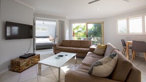 A modern living room with a sofa, coffee table, and TV. There is a balcony with a view of greenery. - Coolum Beach Getaway Resort (Coolum Beach)