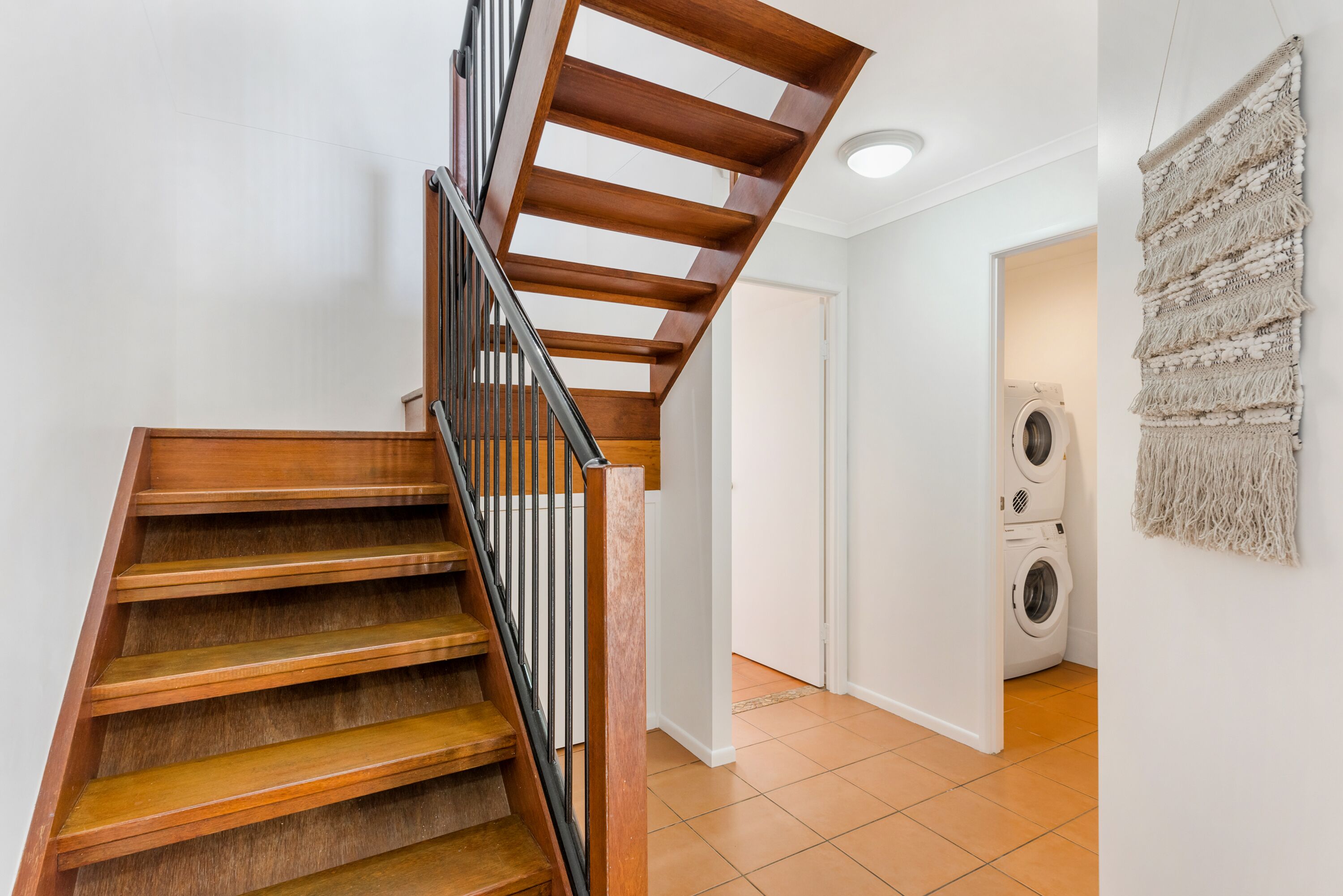Un escalier en bois avec une rampe noire mène à une buanderie équipée de deux machines à laver.