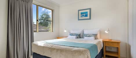 Ein Schlafzimmer mit einem Bett, einem Nachttisch, einem Deckenventilator, einem Fenster mit Vorhängen und einem gerahmten Bild an der Wand.