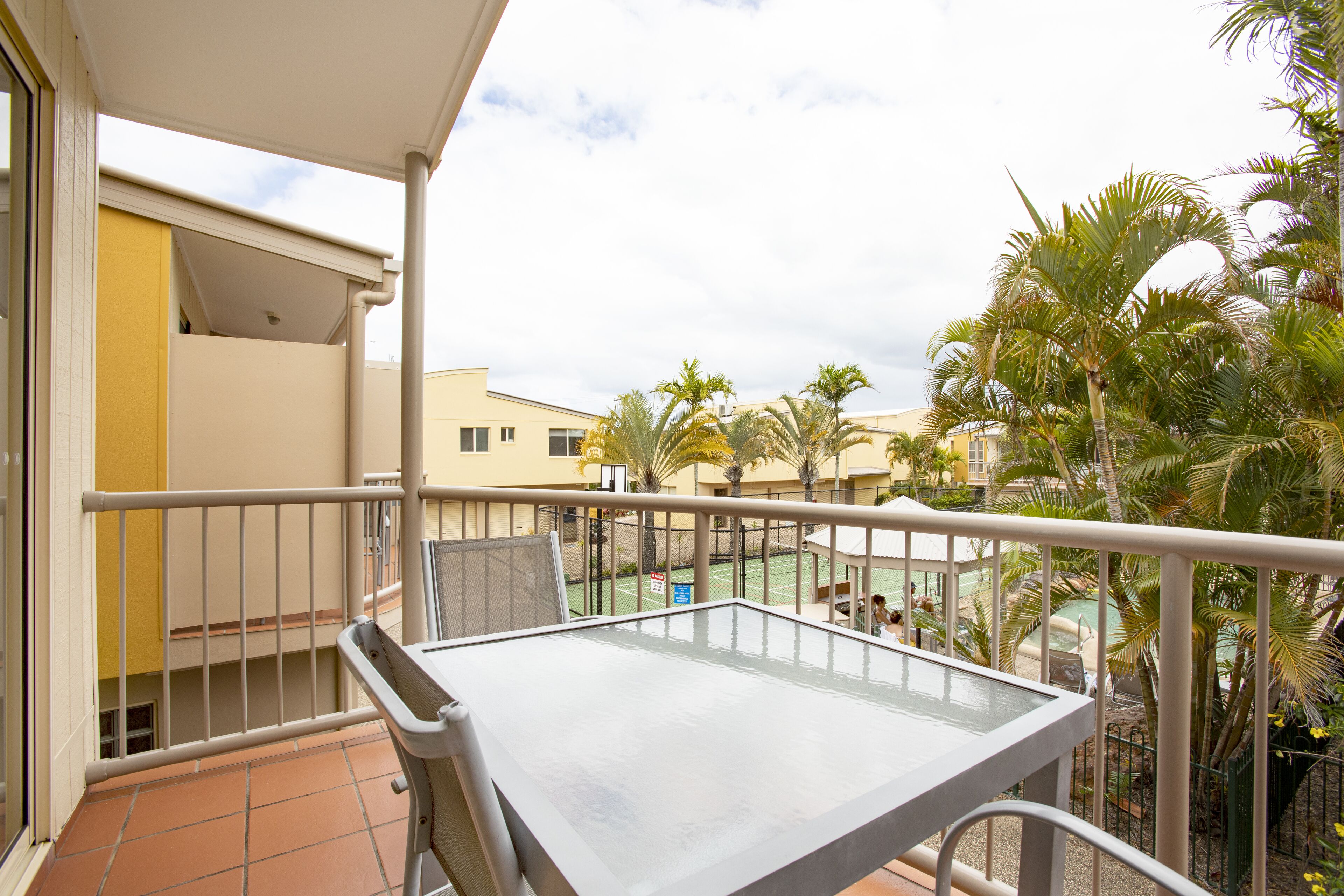 Un balcon avec une table et des chaises, donnant sur un court de tennis et des palmiers.