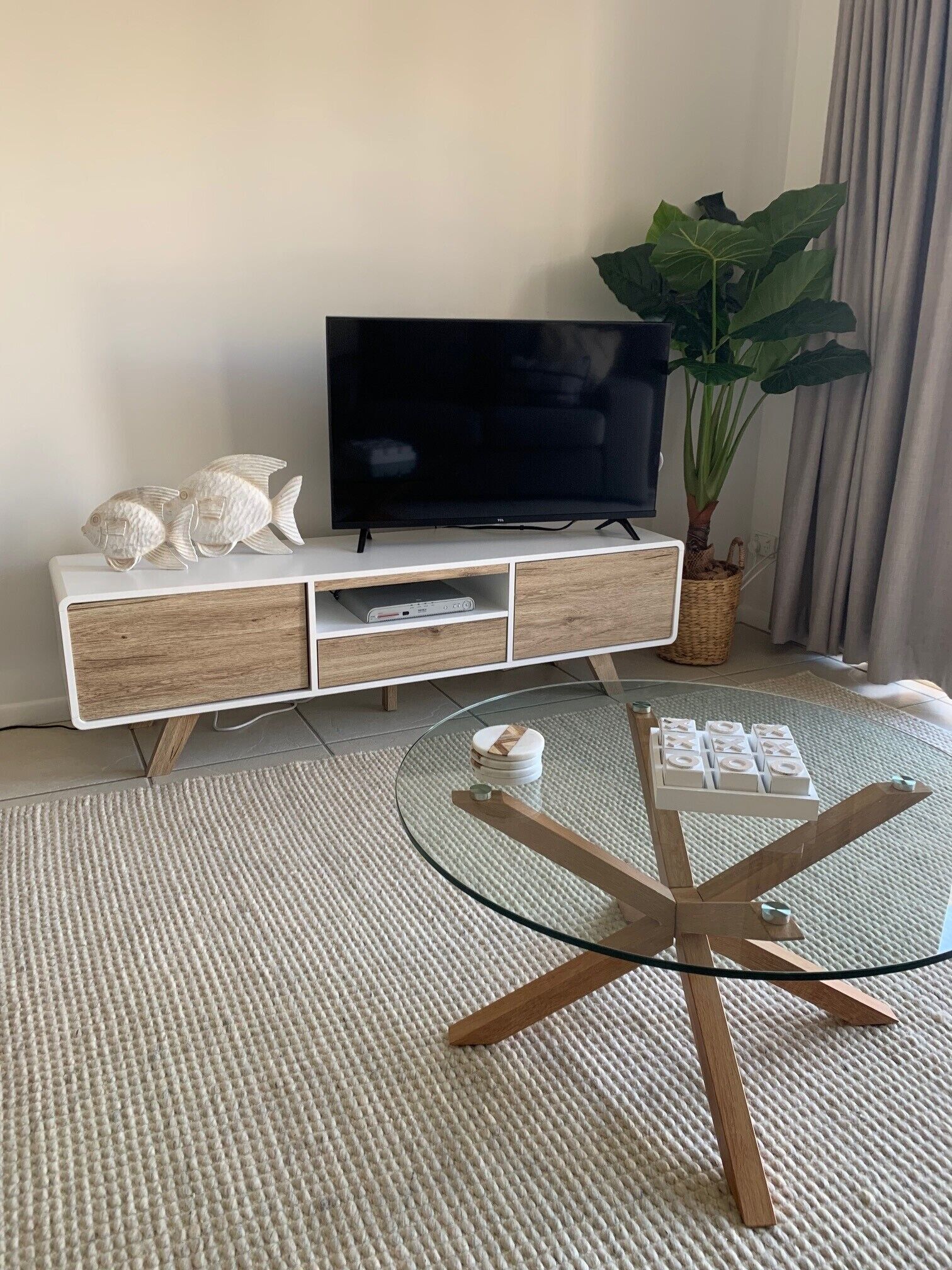 A modern living room with a flat-screen TV on a white and wood console, a glass coffee table with wooden legs, a large plant, and a woven basket.