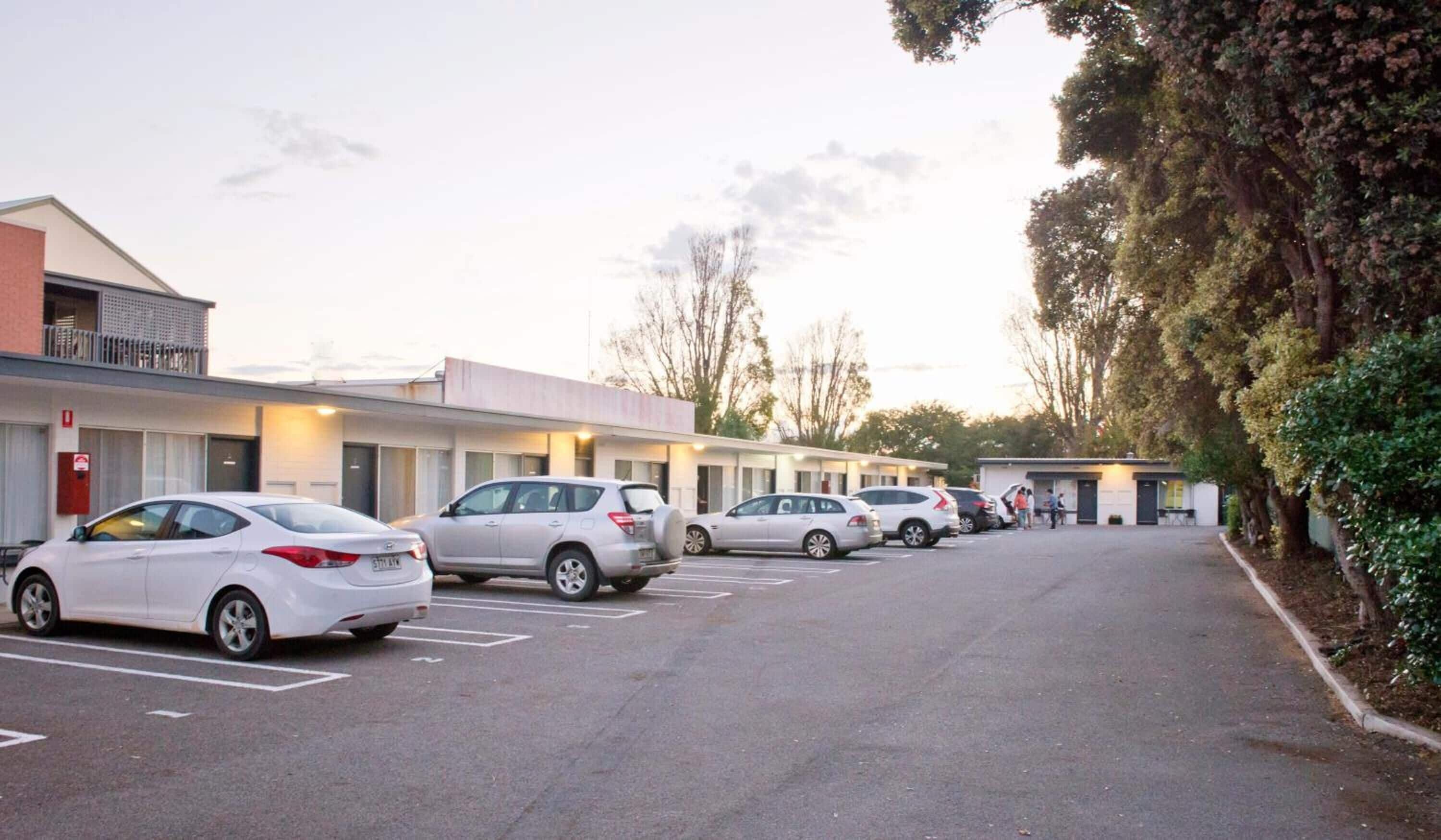 A row of parked cars in front of a motel with a single-story structure and a balcony on the left.