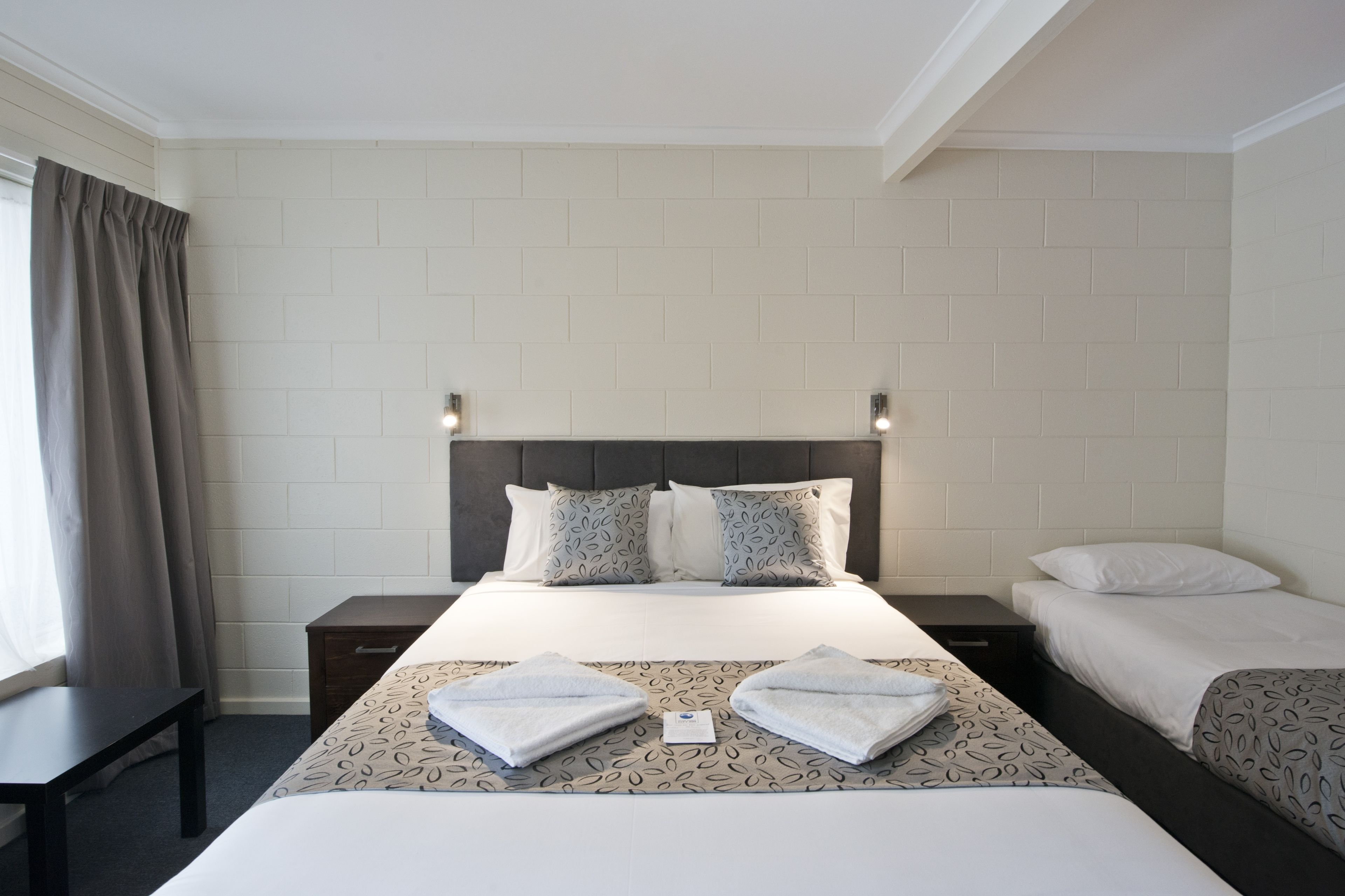 Una habitación de hotel con una cama grande, dos mesitas de noche, un ventanal con cortinas y una pared con diseño de ladrillo.